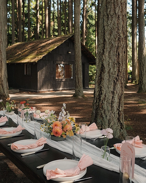Outdoor reception table set with white plates and pink napkins on a black wooden table, with bud vase florals in a forest cabin setting