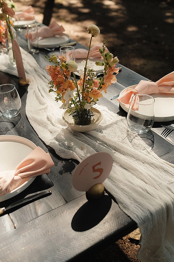 Reception tablescape with peach and white bud vase centerpiece, knotted peach napkins, clear glassware, and white runner under tree shade outdoors
