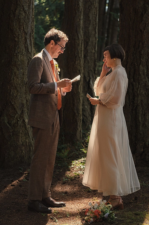Wedding vows as couple reading vows from cards, bride in long-sleeve dress and groom in suit on a sunlit forest path among tall trees
