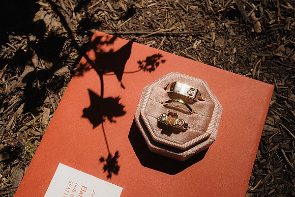 Wedding rings in a wedding ring flat lay with gold and gemstone bands in a velvet ring box atop an invitation and envelope on mulch ground