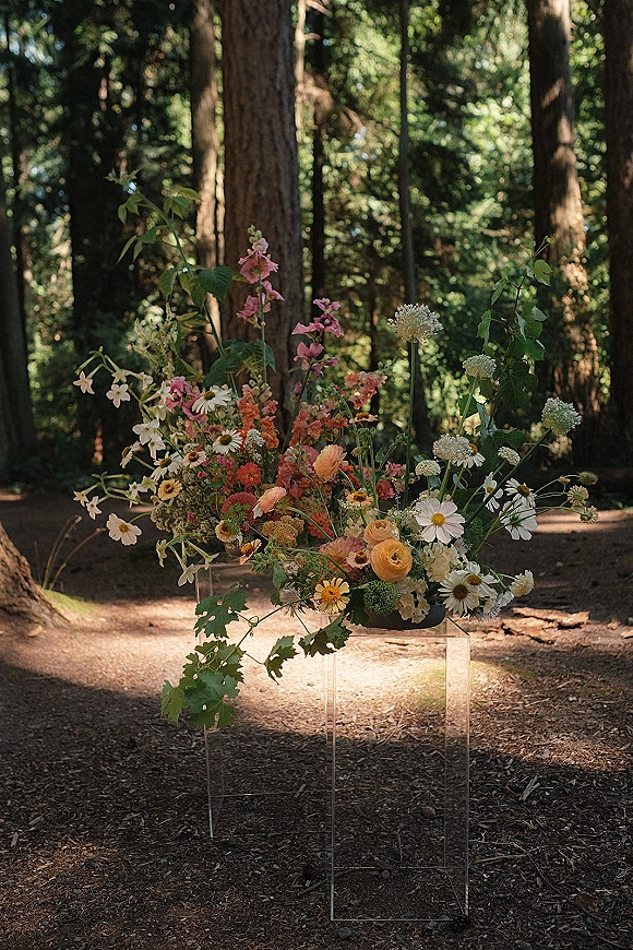 Wedding floral arrangement wildflower wedding centerpiece with white daisies, peach ranunculus and pink blooms on an acrylic pedestal in woodland light