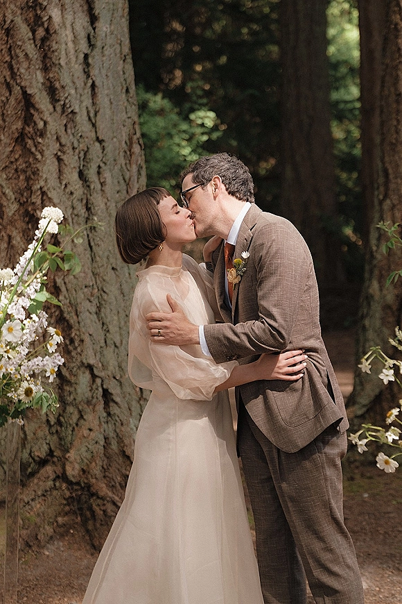 Wedding kiss portrait of bride and groom sharing an outdoor wedding kiss in a redwood forest, his boutonniere against her dress and earrings