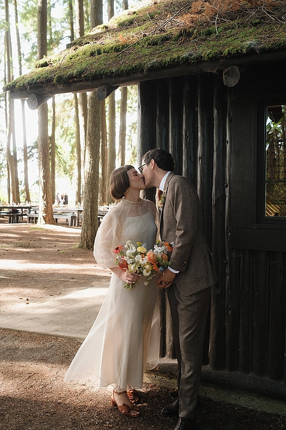 Wedding kiss portrait of bride and groom kiss by a rustic moss-roof cabin, bride holding bouquet and wearing a veil and long sleeves
