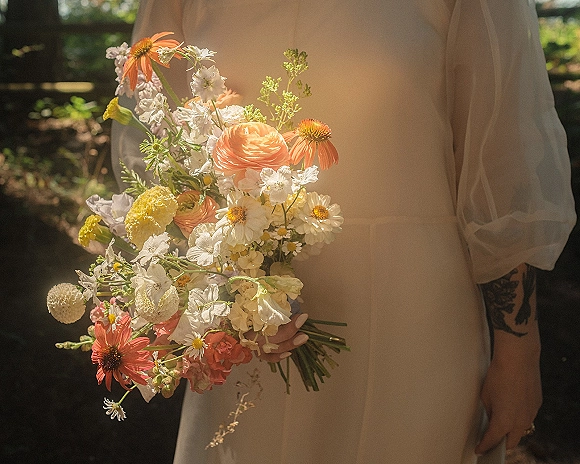Bridal bouquet of wildflowers with peach ranunculus, orange coneflowers, and white daisies in bride’s hands, sunlit garden backdrop