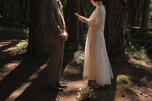 Wedding vows as the bride reads from a vow book and the groom listens, holding a bouquet on a dirt path in dappled redwood forest light