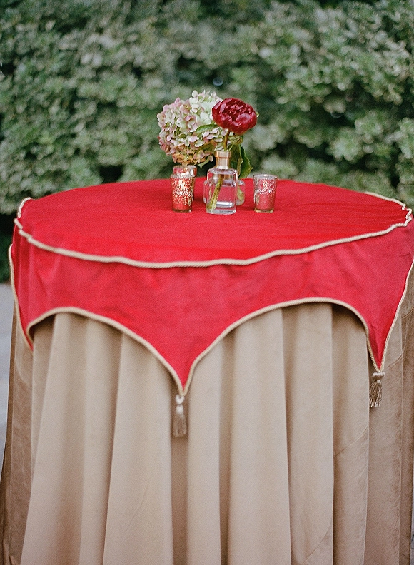 Cocktail table decor with red table topper, beige linen, hydrangea and ranunculus centerpiece, and mercury glass votive candles by a green hedge wall
