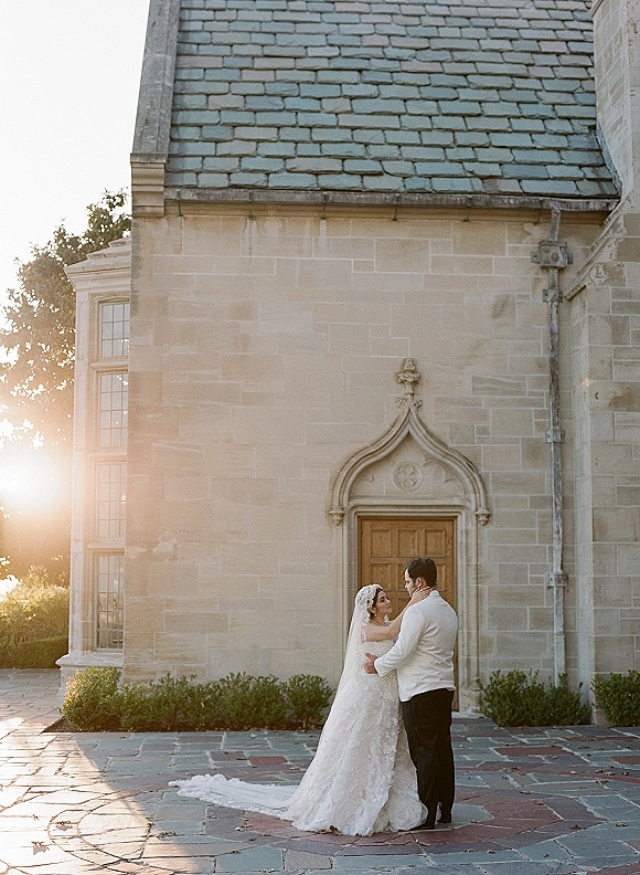 Couple portrait of bride and groom embrace, bride touching his face as sunset light glows by a stone manor arched doorway