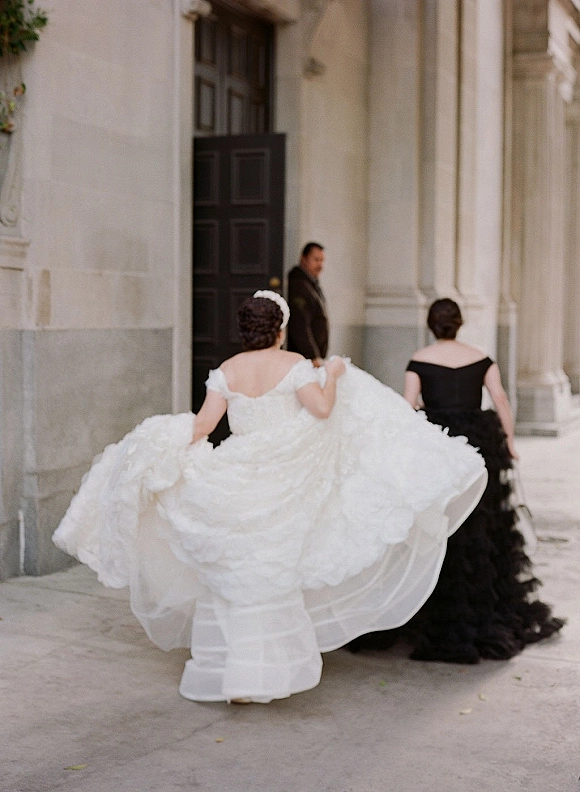 Bride entrance as she walks toward a grand stone doorway, holding her off-the-shoulder gown train while a bridesmaid in black follows behind