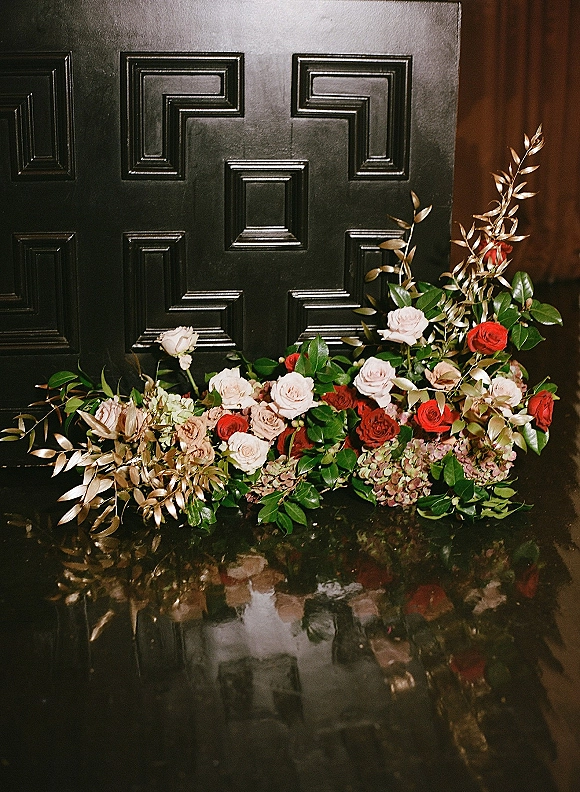 Wedding floral arrangement with red and blush wedding flowers, roses and hydrangea with greenery on a reflective tabletop by a black paneled door
