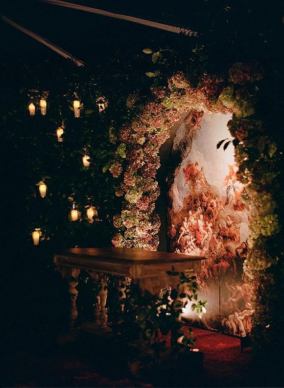 Wedding ceremony backdrop with a floral ceremony arch of hydrangeas and greenery, hanging candles, and mural wall in a dark indoor space