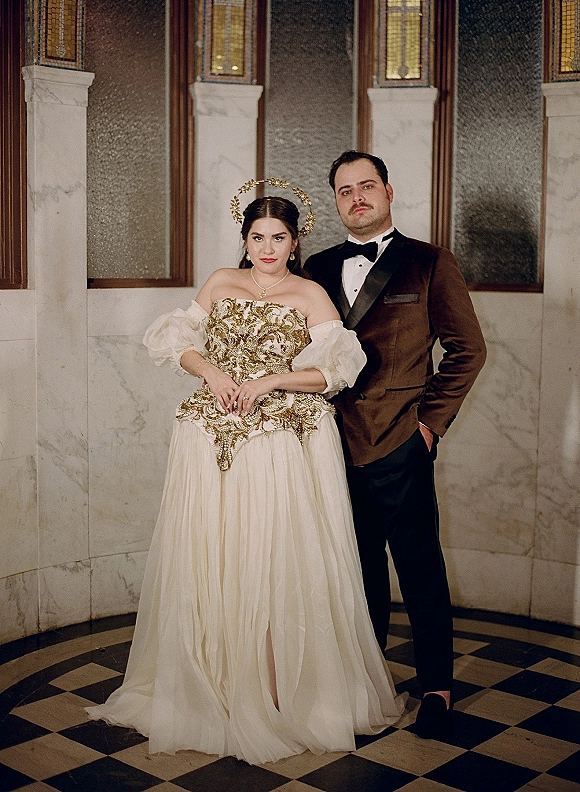 Couple portrait of bride in gold embellished wedding dress with halo headpiece and groom in velvet tuxedo by stained glass windows