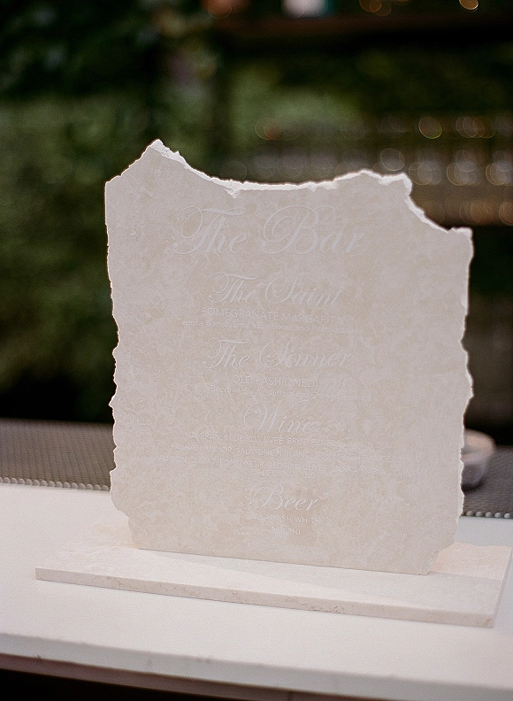 Wedding bar menu with signature cocktail menu in white calligraphy on deckle-edge paper, displayed on a stone base at an outdoor bar with bokeh lights