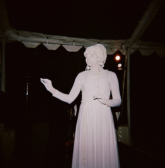 Wedding performance under tent with a performer in a long white dress and knit bonnet, lit by stage lights inside a dark night event tent