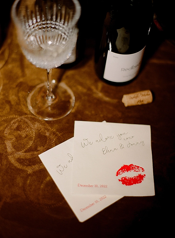 Wedding invitation with envelope and red lipstick kiss mark, calligraphy note beside wine bottle and crystal glass on patterned fabric tablecloth