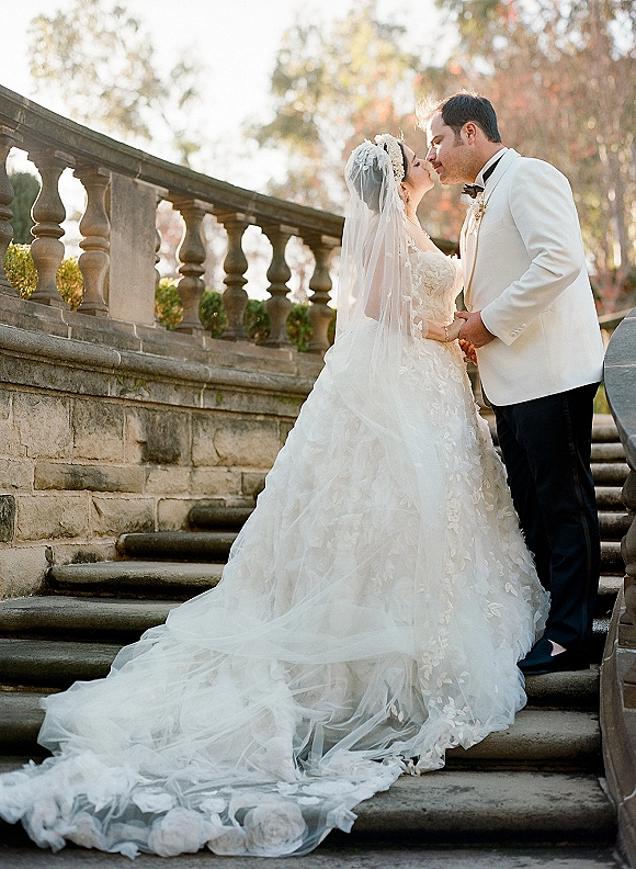 Wedding couple portrait of bride and groom kissing on a stone staircase, her cathedral veil flowing as he wears a white tux and boutonniere