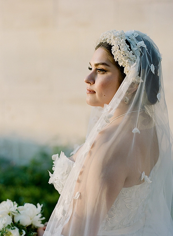 Bridal portrait of a side profile bride in a strapless lace gown with veil and floral headpiece, holding a white bouquet outdoors under sky