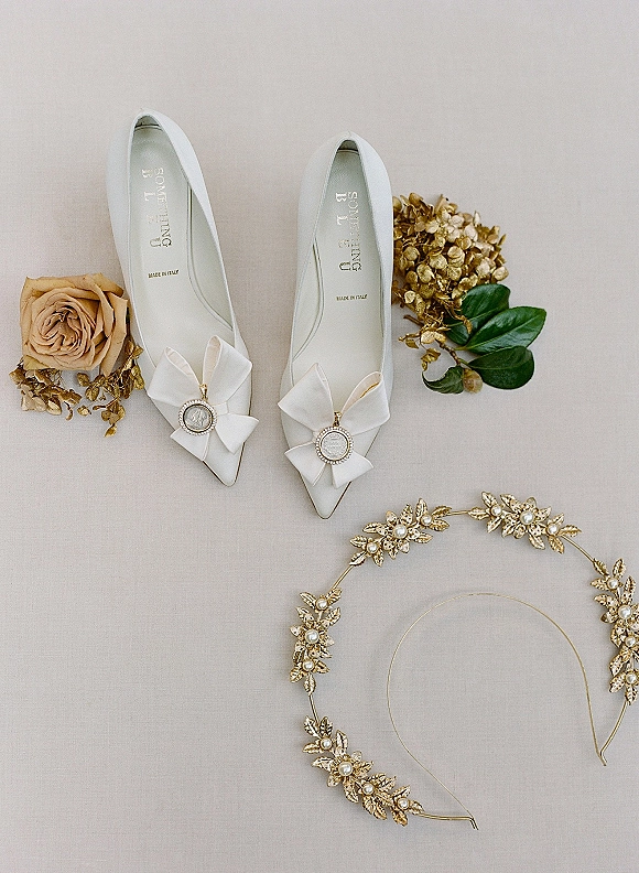 Bridal shoes flatlay of white wedding heels with satin bows and rhinestone clips, styled with dried roses and gold headband on neutral fabric