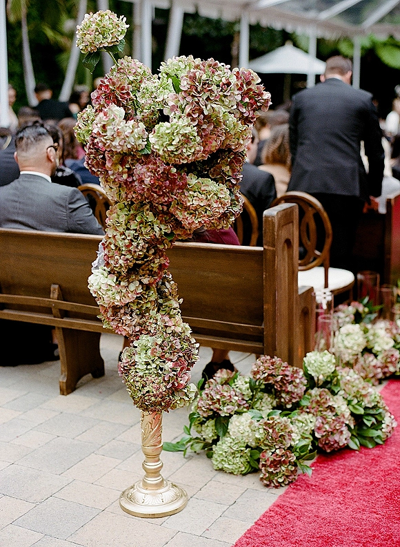 Ceremony aisle decor with hydrangea aisle flowers on a gold pedestal beside wooden pews and a red runner under a canopy outdoors
