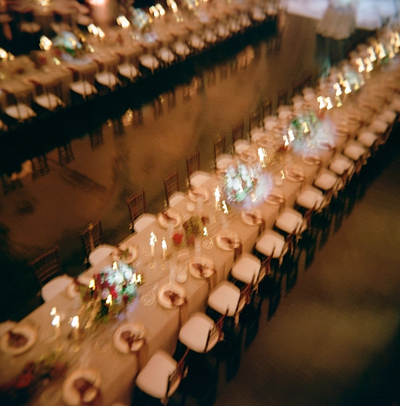 Reception tablescape on a long banquet table with white linens, place settings, glassware, taper candles, and floral greenery in candlelit hall interior