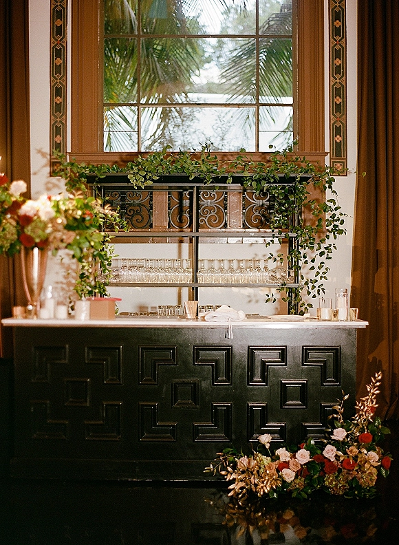 Wedding bar setup with a mobile wedding bar, black front, glassware shelves, and rose greenery garland by an ornate window with palms