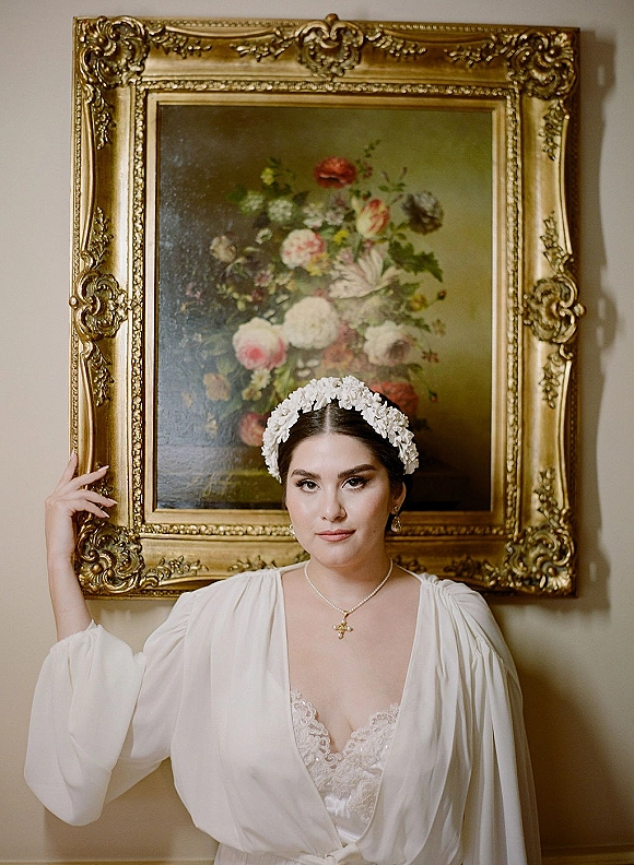 Bridal portrait of a bride in a robe with white floral headpiece, lace camisole, and cross necklace before an ornate gold frame and painting