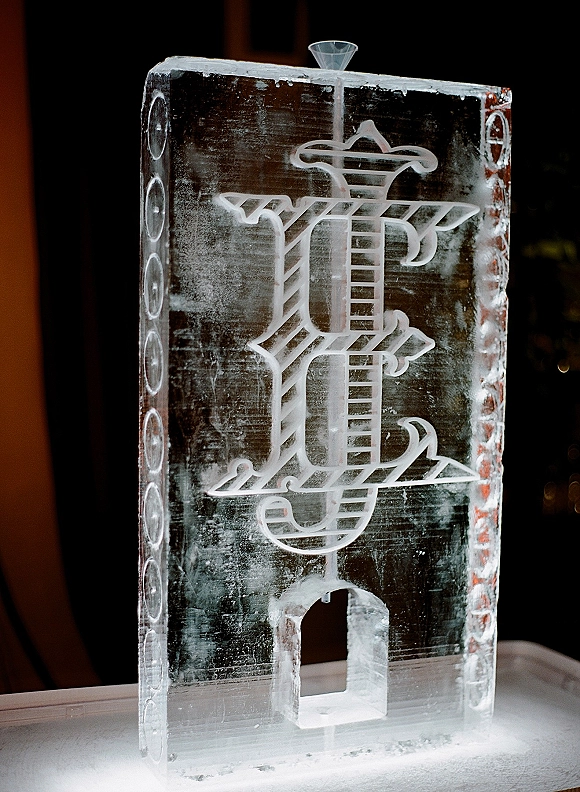 Ice sculpture wedding ice sculpture with carved monogram and funnel ice luge on a tabletop against a dark indoor reception backdrop