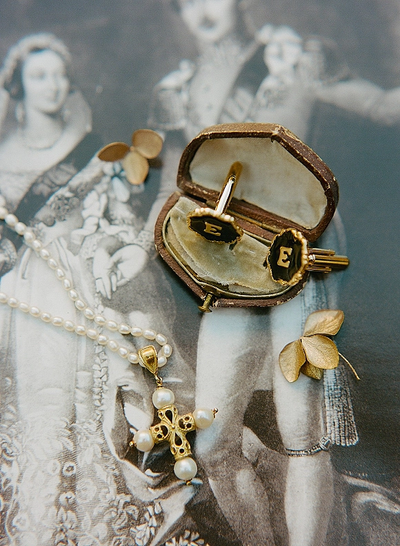 Wedding jewelry flatlay with pearl cross necklace, monogram cufflinks, and velvet ring box on a vintage black-and-white portrait print backdrop