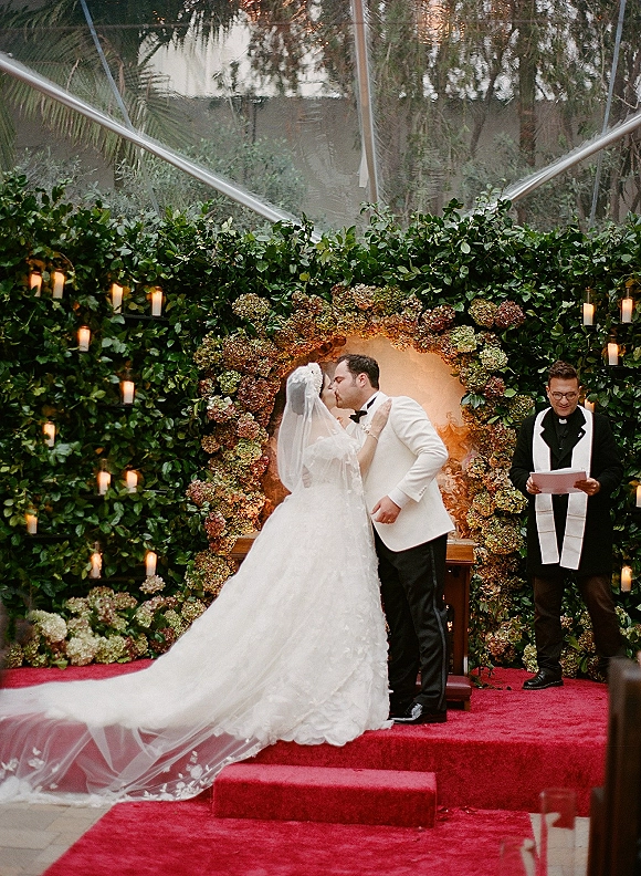 Wedding kiss under a hydrangea floral arch with cathedral veil and long train, groom in white tuxedo on red carpet garden altar