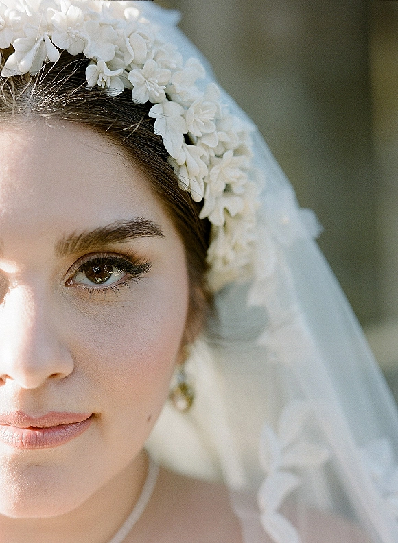 Bridal portrait of a bride in a floral headpiece and veil, wearing drop earrings, in soft bokeh outdoor greenery backdrop