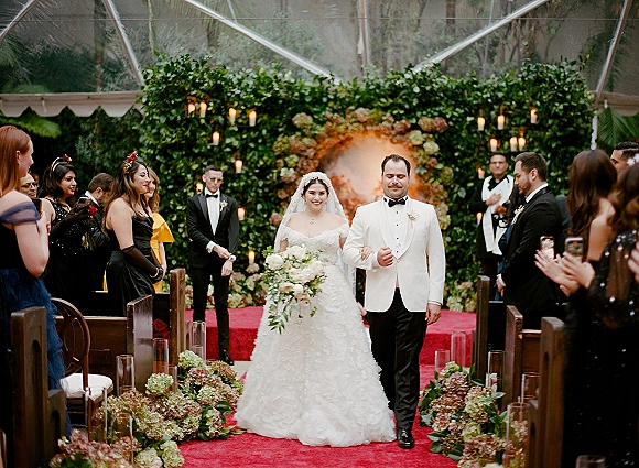 Wedding recessional as bride and groom walk the aisle, cathedral veil and bouquet, under a tent canopy with guests and candles on a red runner