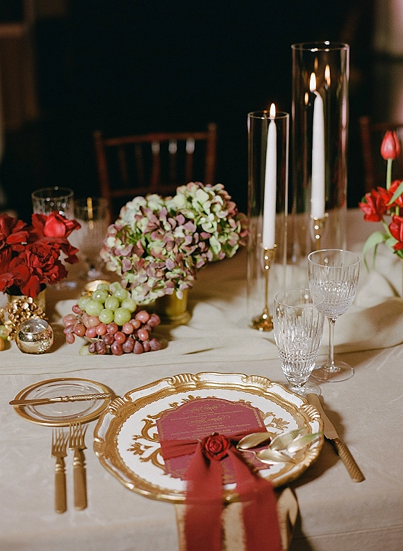 Reception tablescape with a wedding place setting, gold-rim plate, crystal glassware, red menu card and candlelit red florals in a dark interior