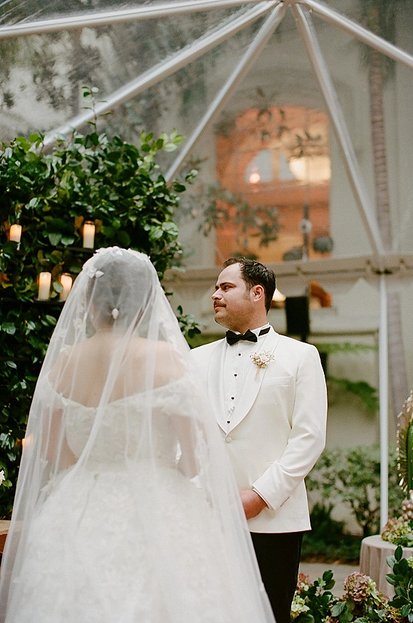 Ceremony moment as bride in ball gown and cathedral veil approaches groom in white tux under greenery arch with candles in glass greenhouse