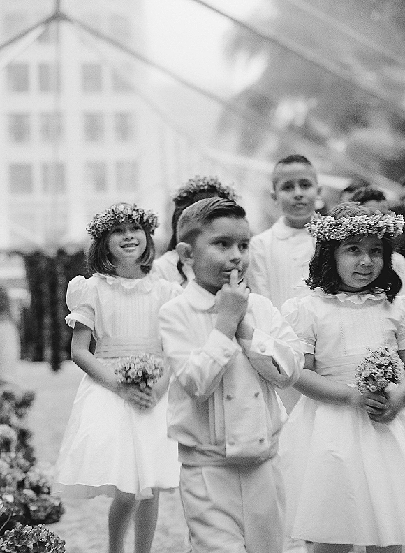 Flower girl and ring bearer walking the aisle, she wears a baby’s breath crown and bouquet, he in a white suit with suspenders under a tent canopy