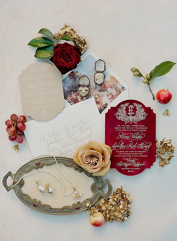 Invitation suite with burgundy wedding invitations on white fabric, styled with wax seal, rings, pearl jewelry, roses, grapes, and silver tray