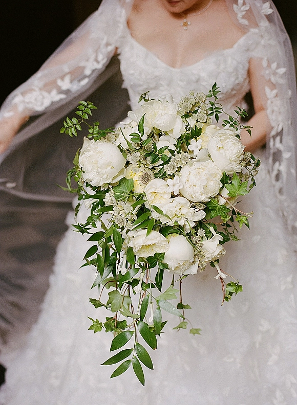 Bridal bouquet of white peonies with trailing greenery, held against lace wedding dress sleeves and veil in a dark interior setting