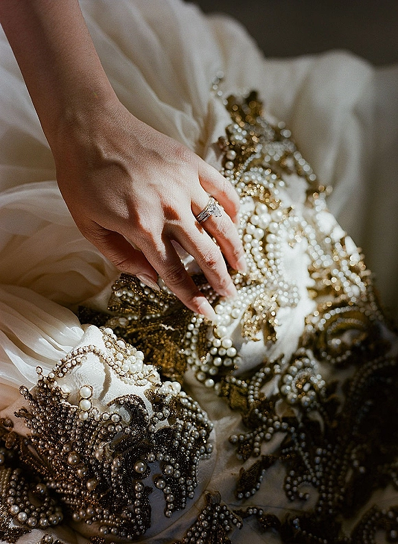 Engagement ring close-up, diamond engagement ring on hand against pearl-beaded wedding dress with gold embroidery on white fabric shadows