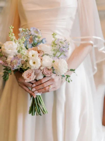 Bridal bouquet of pastel wedding bouquet roses and white blooms with greenery, held against a neutral indoor wall, ribbon sash and rings visible