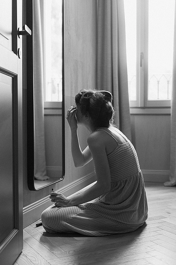 Bridal getting ready as bride applies makeup in a spaghetti strap slip dress, lipstick in hand, by a floor mirror in window light