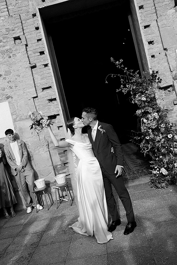 Wedding kiss as bride holds bouquet up and embraces groom in suit, long veil trailing by a stone doorway with confetti on paving