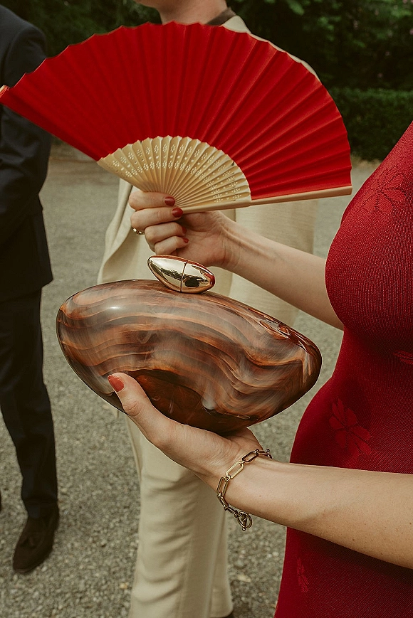 Wedding guest accessories with a red folding fan and marbled clutch, gold clasp and bracelet, red dress and manicure on outdoor pavement