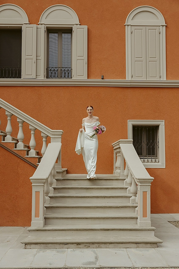 Bridal portrait of a bride in an off-the-shoulder wedding dress holding bouquet and veil on an outdoor stone staircase by arched windows