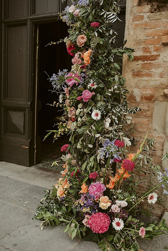 Floral ceremony arch with asymmetrical wedding arch flowers in roses, dahlias, and greenery framing a stone doorway and wooden door