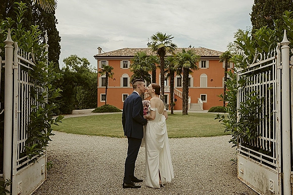 Wedding kiss portrait of bride and groom kissing at a wrought iron gate, bride holding a bouquet before a villa with palm trees behind