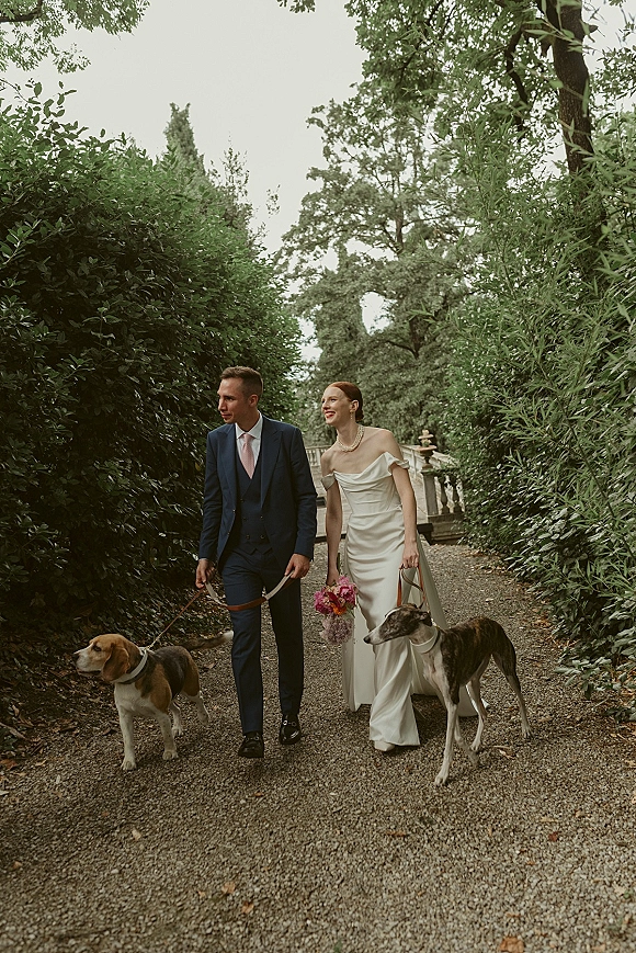 Couple portrait of bride and groom walking with dogs on leashes, bride in veil holding pink bouquet, along a garden path by a fountain