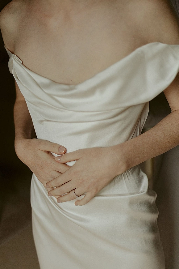 Wedding dress close-up showing an off the shoulder wedding dress with draped satin bodice and bride’s hand with wedding band in neutral indoor light