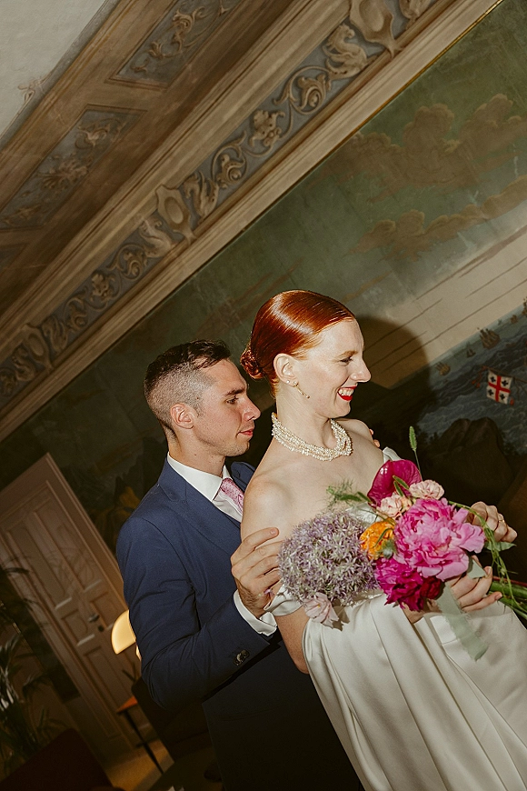 Couple portrait of groom hugging bride holding a bright bouquet, her strapless dress and pearl necklace before a painted mural wall