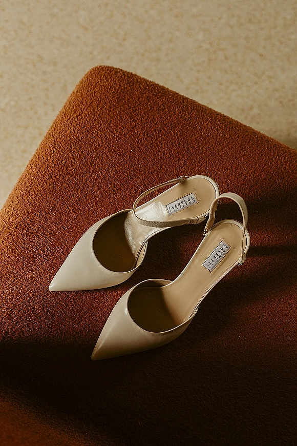 Bridal shoes, ivory wedding heels styled as pointed toe slingback pumps resting on a rust velvet cushion on a neutral floor