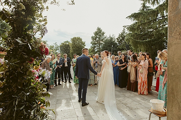 Wedding ceremony moment as bride and groom holding hands walk through cheering guests on an outdoor terrace, confetti falling around them