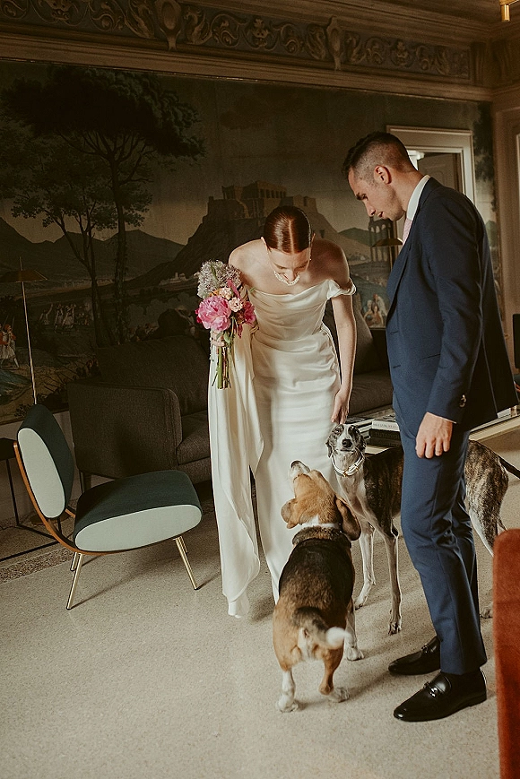 Wedding couple with dogs cuddling beside a mural wall as the bride pets a dog, holding a pink peony bouquet in a satin gown