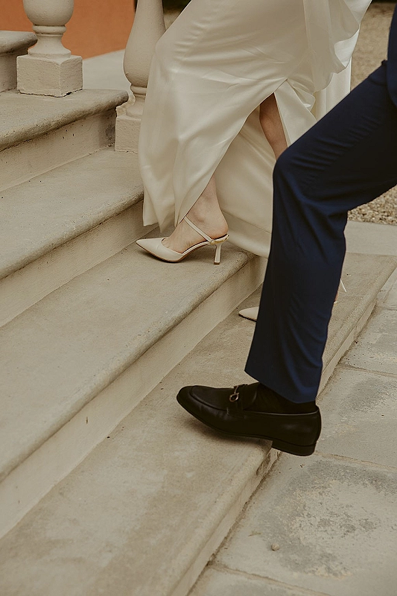 Wedding shoe detail of bride in bridal slingback heels beside groom’s dress shoes, satin dress hem brushing stone steps on outdoor staircase